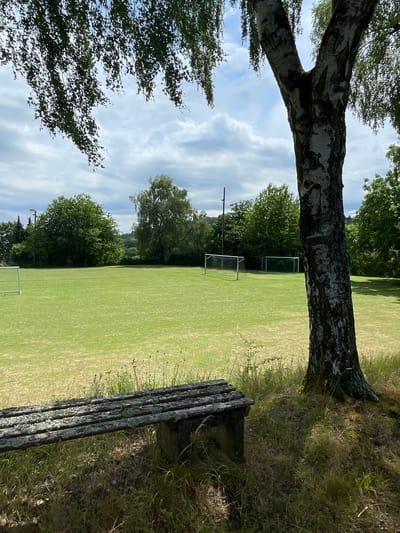 An outdoor, eye-level, medium shot captures a weathered wooden bench positioned in the foreground, partially shaded by the trunk of a large birch tree. The bench is situated on a grassy verge next to a well-maintained green sports field. In the middle ground, the expansive green field is dotted with two white soccer goals, one closer and larger than the other further away. A line of trees forms a dense backdrop across the field's horizon, with a few scattered structures, possibly lights or poles, visible behind them. The sky above is partly cloudy, with dramatic white clouds against a pale blue backdrop. The image appears to be taken on a sunny day. This scene depicts a public sports facility, likely in the city of Olberode, Germany, offering a space for recreation and sports.
