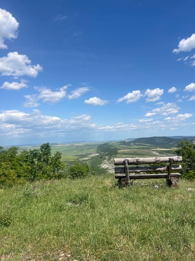 A vertical-format photograph taken in Vetrino, Bulgaria, displays a landscape with a wooden bench on a grassy edge. The bench, constructed from logs, faces out towards a vista of rolling hills and valleys under a blue sky dotted with cumulus clouds. Lush green grass and sporadic bushes dominate the immediate foreground, while the background transitions from green fields to distant mountains under a hazy horizon.