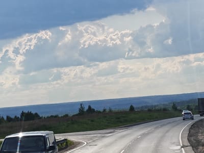 This is an outdoor, wide-angle, eye-level shot of a rural landscape. The sky is dominated by dramatic, white, puffy clouds against a blue sky. In the foreground, a dark-colored minivan is parked on the left side of the frame, its windshield reflecting the sky. To the right, a paved road curves into the distance, with lane markings visible. A light-colored car is driving away on the road, and further in the distance, a large truck is partially visible. The road is bordered by green grass and sparse trees. Beyond the immediate roadside, rolling hills covered in trees and vegetation stretch into the background, fading into a hazy blue expanse under the cloudy sky. The scene appears to be set in a rural area, likely in or near the city of 50 Лет Победы Снт, Russia, given the visual cues of open spaces, trees, and a highway. There are no visible people.