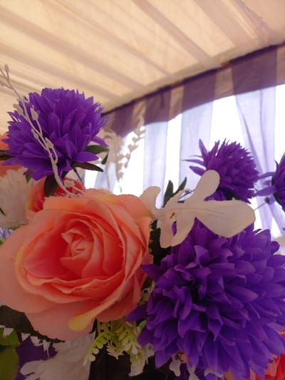The image displays an upward-angled view of an artificial floral arrangement, captured within a setting in Ohafia, Nigeria. The foreground is dominated by a cluster of artificial flowers, primarily featuring several vibrant purple chrysanthemum-like blooms and a prominent, large peach-colored artificial rose. White artificial twig-like elements and smaller green and white artificial foliage are interspersed throughout the arrangement. In the upper background, a light-colored, possibly beige or cream-colored, fabric canopy or tent ceiling is visible, showing linear folds or seams. Below this canopy and behind the flowers, bright light filters through sheer purple curtains, which appear to cover a window or opening, creating a softly illuminated backdrop. No individuals or actions are depicted in the scene.