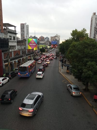 The image provides an elevated view of a congested urban street in Ramos Mejía, Argentina, under an overcast sky. The dark asphalt road surface exhibits some reflectivity, suggesting dampness. Vehicular traffic is heavy, with multiple cars stopped or moving slowly in a traffic jam extending into the distance. A blue and white public bus (Line 172) is centrally positioned on the left side of the street, moving away from the viewer. In the immediate foreground, a dark grey Audi SUV also travels away from the viewer, with a black sedan to its left. A silver sedan is parked parallel to the curb on the right side of the street. On the left, multi-story buildings line the street, featuring commercial storefronts and a large, colorful billboard displaying "PACHOS." On the right, a sidewalk lined with green-leafed trees runs alongside the road, featuring a blue bus stop shelter. A single individual in a red shirt and dark pants is visible walking on the far right sidewalk, moving away from the camera. Several other indistinct pedestrians are observed on the sidewalks further down the street.