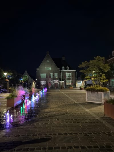 The image depicts a night scene in the city of Boxtel, Netherlands, specifically at the Markt square. In the foreground, a row of colorful water fountains illuminated by LED lights creates a vibrant spectacle, their reflections shimmering on the wet cobblestone pavement. Planters filled with grass and ornamental plants line the fountains, adding a touch of nature to the urban landscape.
Dominating the background is a large, brick building with a prominent gabled roof, characteristic of Dutch architecture. Its windows are illuminated, suggesting activity within. To the right of this building, a tree with green foliage stands in a large planter, its branches catching the artificial light. Other buildings and structures are visible in the distance, contributing to the atmosphere of a quiet town square at night. The overall scene is serene and visually appealing, with the interplay of light, water, and architecture creating a captivating ambiance.