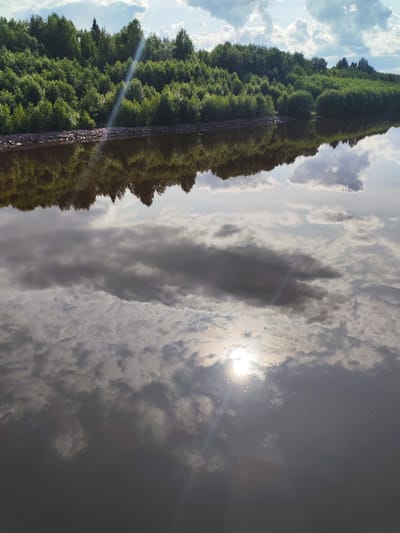 The image shows a body of water with a reflection of a tree-lined shore. The sun is visible in the sky, reflecting brightly on the water. The sky is partly cloudy. The environment appears to be natural, possibly a lake or river in a rural or forested area. There are no people or man-made structures immediately visible in the scene.