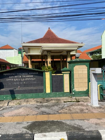The image shows the exterior of the building for the Kecamatan Tulangan, located at latitude -7.48 and longitude 112.645 in the city of Kecamatan Tulangan, Indonesia. The building has a red roof with a white, ornate trim and white columns. The building is surrounded by a green brick wall with a plaque on the front that says, "PEMERINTAH KABUPATEN SIDOARJO KECAMATAN TULANGAN KANTOR KEPALA DESA KEPADANGAN". The building is situated in a residential area with other buildings visible in the background. There are many power lines in the sky above the building. The ground is concrete with a cracked sidewalk in front of the building. There is a large, white, concrete pillar on the right side of the image.