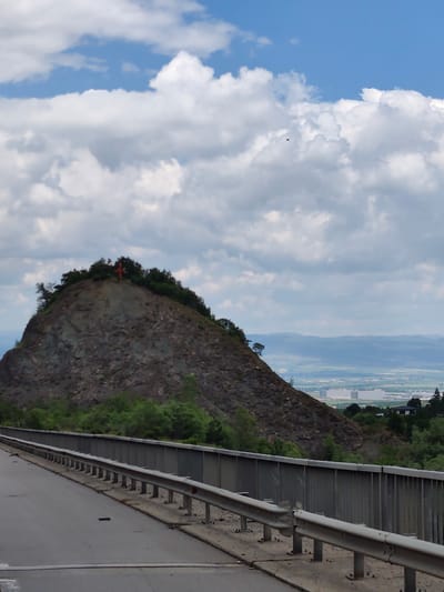 An overcast sky with white and grey clouds hangs above a rugged, tree-covered hill. At the very peak of the hill, a bright red cross is visible, appearing to be a religious monument. The hill is situated in Malo Buchino, Bulgaria. In the foreground, a road with a metal guardrail stretches towards the left side of the frame. Beyond the hill, a hazy landscape unfolds, revealing distant mountains and a sprawling urban or industrial area with large, light-colored buildings. The overall scene suggests a view from a road overlooking a town and its natural surroundings.