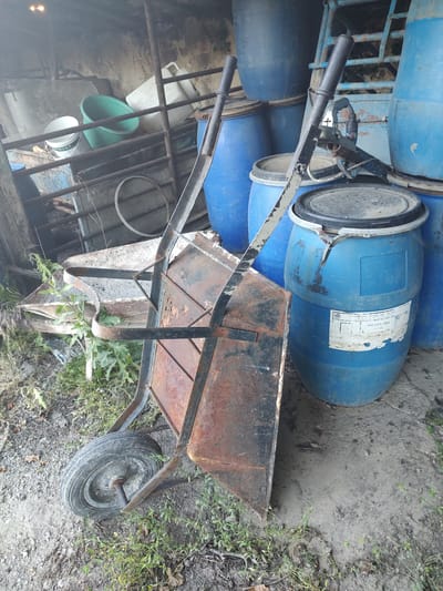 The image shows an outdoor scene, likely in a yard or storage area, with several large blue plastic barrels. In the foreground, a rusted wheelbarrow with a black handle is positioned horizontally across the frame. The wheelbarrow's tray is made of metal and shows signs of wear and tear. A single wheel with a tire is visible on the right side of the wheelbarrow. The ground is a mixture of dirt, gravel, and some sparse green vegetation.
Behind the wheelbarrow, several of the blue barrels are stacked and leaning against each other. One barrel has a white label with some text on it, but the text is mostly illegible. The barrels appear to be storage containers, possibly for liquids. To the left and behind the barrels, there are some metal bars, suggesting a structure like a fence or shelving. Further in the background, a glimpse of a brick wall and more barrels can be seen, along with some green and white containers. The overall impression is one of a storage or work area, possibly a bit unkempt.
There are no people visible in the image. The lighting suggests it could be daytime, possibly overcast given the lack of strong shadows. The scene appears to be located in a rural or semi-rural setting, given the ground cover and the presence of the barrels and wheelbarrow, which are commonly found in such environments. The visible text on the barrel label is too blurry to be read.