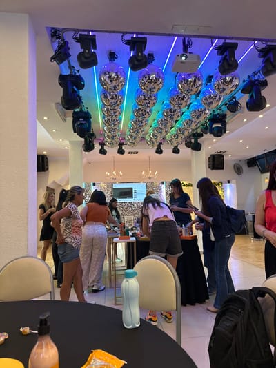 The image captures an indoor event space, characterized by a lively and modern aesthetic. On the left side, a prominent white wall is adorned with multiple rows of glittering disco balls, creating a dynamic visual effect. These disco balls are illuminated from behind by horizontal strips of LED lighting, which transition through vibrant colors including blue, green, and hints of pink and purple, suggesting a mood-setting or party atmosphere. Mounted on the same wall are several black stage lighting fixtures, resembling moving head lights, positioned above and below the disco ball rows. A white projector is also visible on this wall, situated among the lighting equipment.
On the right side of the image, a group of women is gathered, mostly standing and engaging in conversation or looking at various items. In the upper right, a woman in a red top is partially visible. Below her, a woman wearing a dark blue jacket and blue jeans stands with a black backpack, looking downwards, possibly at a table in front of her. Further into the scene, another woman in a light-colored top and dark shorts leans over a light-colored chair, with her colorful sneakers (featuring orange, yellow, and white elements) clearly visible. Several other women are distributed throughout the mid-ground, dressed in casual attire, some with their backs to the camera, others in profile. One woman wears an orange knit top and light-colored patterned pants, while another sports a patterned top and blue shorts.
In the immediate foreground, a black round table holds several items: a light blue Tupperware water bottle with a turquoise lid, a clear plastic bag containing orange-colored items (possibly snacks or candy), a pump dispenser bottle filled with an orange liquid, and a scattering of small, colorful candies. Light-colored, possibly beige or cream, chairs are positioned around this table. In the background, beyond the immediate group of people, more activity is suggested with additional individuals and tables. A screen displaying what appears to be a video player interface on a laptop or monitor can be faintly seen. Elegant chandelier-style light fixtures hang from the ceiling in the distance. The overall setting suggests a social gathering, possibly a party or a promotional event, marked by its vibrant lighting and festive decorations. No explicit text is clearly legible in the image.