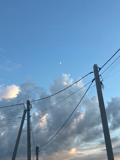 The image depicts a sky scene with power lines and poles in the foreground, against a backdrop of clouds and a crescent moon. The sky is a gradient of blue, with lighter shades near the horizon where clouds are illuminated by sunlight. The clouds are varied in texture and density, ranging from wispy streaks to more solid, billowing formations. A thin crescent moon is visible in the upper right quadrant of the sky. Two prominent concrete power poles, with multiple wires suspended between them, occupy the right and left sides of the frame. The poles appear weathered and utilitarian, with various attachments and coiled wires visible on the pole on the right. A smaller, less distinct power pole is visible in the lower right portion of the image. The overall impression is one of an outdoor scene, possibly during twilight or early evening, with a focus on the infrastructure against a natural sky. There are no people or discernible actions occurring in the image.