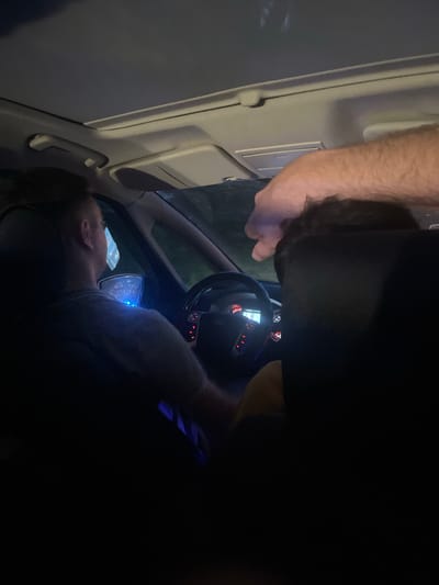 The image depicts the interior of a car at night. A person is seen from the neck up, lying down, with their head resting on something or someone. Another person's arm and part of their head are visible entering the frame from the top. The dashboard and steering wheel of the car are illuminated by the interior lights and possibly the screen of a phone mounted on the dashboard, which displays blue light. The overall lighting suggests it is nighttime, and the location is identified as Beloci, Moldova. There are no discernible weather conditions or strong emotional cues. No visible text is present in the image.