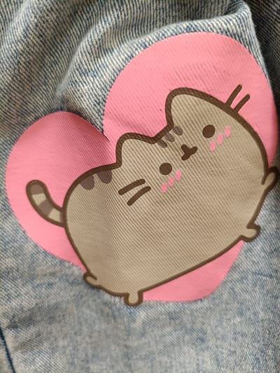 The image shows a close-up of a pink heart with a cartoon cat character, Pusheen, printed on it. The print is on blue denim fabric, likely from an article of clothing. The cat is gray with dark brown outlines and details, including its eyes, nose, mouth, and whiskers. It has small pink circles on its cheeks and two small pink lines above its nose. The heart is a bright pink and has a textured surface. The denim fabric shows the characteristic diagonal weave and some creasing, indicating it is flexible and likely worn. The scene is neutral, with no discernible background elements or environmental context. The image does not provide any information that can be used to determine its location within the city of Koszalin, Poland.