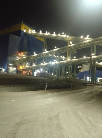 The image captures an industrial facility at night, likely a mining or processing plant, in the vicinity of Қопа ауылдық округі, Kazakhstan. The scene is illuminated by numerous artificial lights, creating a stark contrast against the dark sky. Dominating the frame are complex structures of conveyor belts and support beams, crisscrossing at various levels. These belts are lined with lights, casting a bright glow along their paths. In the background, a large industrial building is visible, distinguished by its blue and yellow facade. The ground in the foreground appears to be unpaved, with a rough, gravelly texture, possibly a road or a work area. The overall impression is one of a functional, operational industrial site at night. No people or specific actions are discernible.