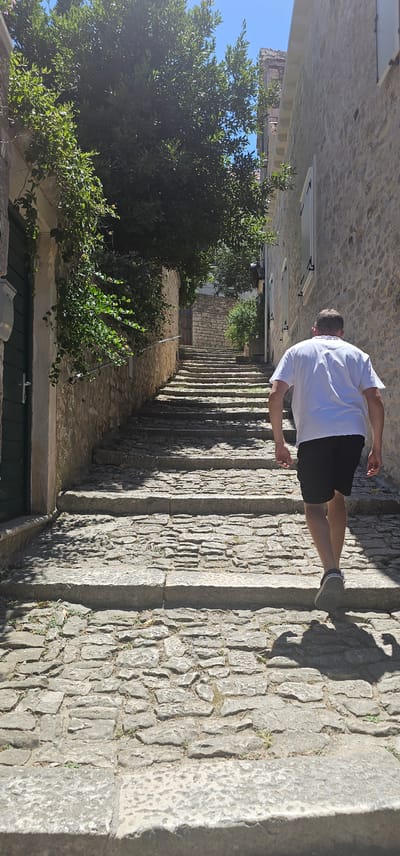 A man walks up a stone stairway in a European city, likely Općina Pučišća, Croatia, given the architecture. The man, wearing a white t-shirt and black shorts, is in the middle of the frame, his back to the camera, ascending the worn steps. The stairway is flanked by stone buildings, one with green shutters, and lush vegetation drapes over a wall on the left. Sunlight casts sharp shadows across the uneven cobblestone steps, indicating a bright, sunny day. The overall scene evokes a sense of historic urban exploration.