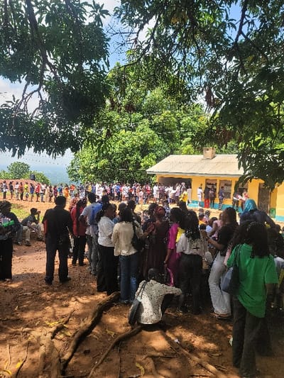 A large group of people are gathered outside a building in Awgu, Nigeria, located at latitude: 6.084, longitude: 7.472. The building has a yellow facade and a metal roof. There are several tree branches in the foreground and a large tree in the background. The people are standing in a line, waiting to enter the building. Some people are dressed in brightly coloured clothing. Many of the people in the image are interacting with each other. Some people are standing in line, and some are talking to each other. It is a sunny day with blue skies. The ground is made of dirt and there are tree roots on the ground. The people are likely waiting to enter the building for a service or a meeting.
