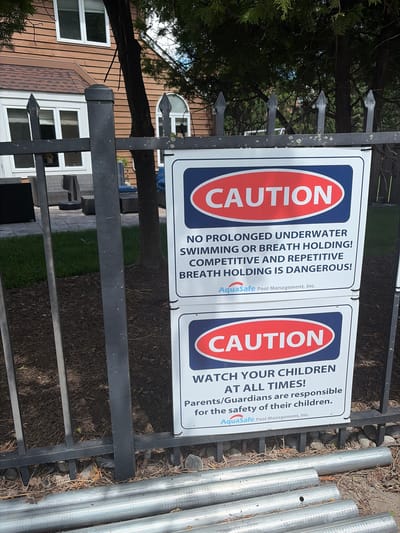 The image captures a scene near a residential area, likely in Edison, United States of America. Focus is on a black metal fence to which two caution signs are affixed. The signs read "CAUTION: NO PROLONGED UNDERWATER SWIMMING OR BREATH HOLDING! COMPETITIVE AND REPETITIVE BREATH HOLDING IS DANGEROUS!" and "CAUTION: WATCH YOUR CHILDREN AT ALL TIMES! Parents/Guardians are responsible for the safety of their children." Both signs feature the "AquaSafe Pool Management, Inc." logo. To the right of the fence, there are several silver metal pipes or poles laying near the ground. Behind the fence, a house with white-framed windows and light brown siding is partially visible. Trees and foliage are present, suggesting an outdoor, possibly landscaped area. The photograph appears to have been taken during daylight hours.