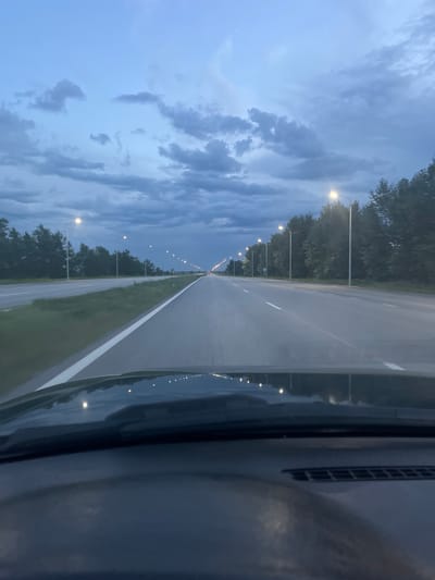 The image shows a view from inside a vehicle driving on a multi-lane highway at dusk. The road is paved and has white lane markings. The sky is overcast with dark, dramatic clouds. Streetlights are illuminated along the side of the road, casting a warm glow. Dense trees line the right side of the highway. The dashboard of the vehicle is visible in the foreground, reflecting some of the lights from the road. The location is identified as Zhdanov rural district, Kazakhstan.