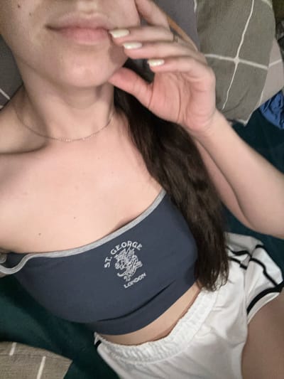 The image shows a close-up of a person's torso, with their hand touching their chin. They are wearing a dark blue strapless top with a graphic design that includes the words "ST. GEORGE LONDON" and an illustration of a lion. Below the top, they are wearing white shorts with black stripes along the sides. The background consists of a dark green surface, possibly a bed or sofa, and a pillow with a grey and white striped pattern. The lighting is soft and diffused, creating a relaxed and intimate atmosphere. The location in Postoyata, Ukraine is not determinable from the image.