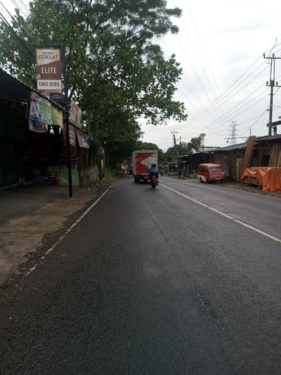 The scene is located in the city of Kamji Na-Yekha, Bhutan. The image presents a street-level view of a bustling roadside on an overcast day. A dark, possibly damp, asphalt road dominates the foreground and midground, featuring a faint white line separating it from a raised, worn concrete sidewalk on the left.
On the left side of the road, a series of simple commercial structures, characterized by corrugated metal roofs and various banners, line the street. A prominent black pole displays a vertical sign with "DIARUM COKLAT ELITE TOKO DERIL" in black text on a white background, featuring a QR code. Numerous colorful posters and banners are affixed to the buildings and the pole. A large tree with abundant green foliage and several light-colored, fruit-like objects (resembling jackfruit) partially obscures these structures.
On the road, a red box truck, likely a delivery vehicle, is moving away from the viewer, followed closely by a person on a black motorcycle. The motorcyclist is wearing a blue jacket and a helmet.
The right side of the road features a dirt shoulder alongside a row of rudimentary wooden structures, some with corrugated roofs, appearing to be workshops or storage sheds. Stacks of raw wooden planks are visible in front of these structures, with some items covered by an orange tarp. A small, red passenger van with a yellow license plate is parked facing away from the viewer. Overhead, numerous utility poles support a dense network of electrical and communication wires. In the distant background, a large metal lattice electricity transmission tower stands against the uniform light-grey, overcast sky.