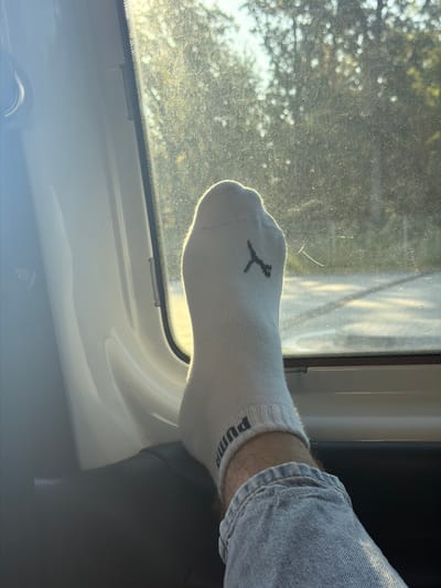 A close-up shot focuses on a person's right foot, clad in a white Puma sock, resting against a car window. The foot is angled upwards, with the sock featuring the black Puma logo and the word "Puma" near the ankle. The individual is wearing light-wash denim jeans, visible below the sock line. The car interior, including the white door panel and dark trim, is partially visible. Through the slightly dusty window, blurred greenery and sunlight suggest an outdoor setting, possibly in the city of Sculeni, Moldova. The sunlight casts a warm glow on the scene, creating lens flare and highlighting the texture of the car window and the sock.