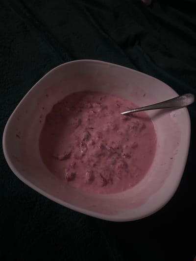 A white bowl filled with a pink, creamy substance containing small, dark chunks is placed on a dark green, textured surface. A silver spoon is resting in the bowl, partially submerged in the mixture. The lighting is dim, casting shadows that obscure some details. There is no indication of a specific location in Postoyata, Ukraine.