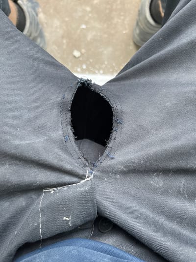 A close-up, overhead shot shows a pair of dark grey pants with a large rip in the fabric. The rip has frayed edges, and there are a few loose blue threads visible around it. The fabric of the pants has a textured, woven appearance. To the left of the rip, the light grey sole of a shoe is visible, suggesting the pants are being worn. The background is out of focus but appears to be a light-colored, possibly concrete or gravel surface, with some white debris scattered around. Further down and to the left, another shoe with a dark sole and grey upper is visible. To the right of the rip, a portion of a darker blue fabric is visible, possibly another garment underneath or part of the pants' lining. There is no visible text in the image. The lighting suggests it might be daytime, possibly outdoors, given the texture and color of the background. The focus is entirely on the damaged pants.