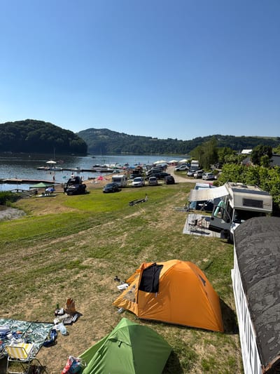 This is a wide-angle, eye-level, outdoor photograph taken on a clear, sunny day at a campsite near a lake. The foreground shows a patch of dry, brown grass with a green tent and an orange tent set up. Beside the green tent, there is a checkered blanket with various items, including a folding chair, clothing, and bottles. Next to the orange tent, there are shoes and a bag. Further back, the terrain transitions to greener grass, where several parked cars, campervans, and a caravan are visible. A road or path runs through the campsite, connecting the vehicles.

Beyond the campsite, a calm lake stretches towards the horizon. Several piers and docks extend into the water, with people swimming and relaxing on the shore. A few boats and sailboats are present on the lake. The background is dominated by lush, tree-covered hills under a clear blue sky. The overall impression is of a recreational area during a warm season. The location is identified as Gródek nad Dunajcem, Poland.