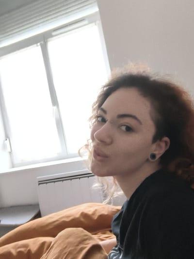 The image depicts a female individual positioned within an indoor setting, specified as the city of La Madeleine, France. The woman, with curly, reddish-brown hair, occupies the right half of the frame, facing slightly left while looking towards the viewer with puckered lips. She wears a dark-colored top. Visible body modifications include a horizontal silver bridge piercing between her eyes, a black ear gauge/plug in her left earlobe, and a small silver stud on the upper helix of her left ear. She appears to be seated or lying on a bed covered with an orange-brown duvet or blanket, which fills the lower left portion of the image. The background on the left is dominated by a brightly lit window with white frames and partially visible horizontal blinds. Below the window, a white wall-mounted radiator is present. To the left of the radiator, a dark, indistinct piece of furniture is partially visible. The overall lighting is bright, originating primarily from the window.