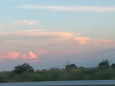 This image captures an outdoor landscape scene at twilight or dawn, presumably in Akhalsopeli, Georgia. The sky dominates the upper two-thirds of the frame, transitioning from a pale blue at the zenith to vibrant pink and orange hues at lower altitudes, indicative of a low sun angle. Large, irregularly shaped clouds are illuminated from beneath, displaying strong pink and orange coloration, with some softer white and grey clouds present. Below the vibrant cloud formation, the sky fades into a muted purplish-grey haze towards the horizon.
The midground features a band of dark green vegetation, including dense trees and bushes, which exhibits slight motion blur. A large, leafy tree is prominently positioned in the left-center. Two slender black streetlights, each with a single lamp, extend vertically from the ground, one on the left and another further right. Faint, indistinct structures or fences are visible along the distant horizon line. In the far background, hazy outlines of hills or mountains are discernible, blending with the atmospheric conditions. A blurry, light-colored horizontal bar occupies the bottom edge of the frame, consistent with the interior of a vehicle from which the photograph was taken. No people or specific human activities are depicted.