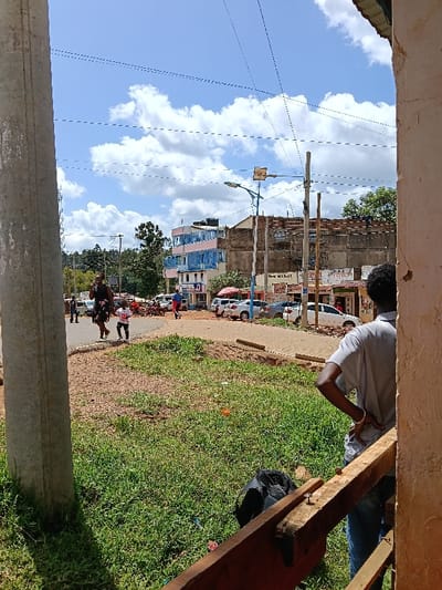 The image depicts an outdoor urban scene in Boochi Tendere ward, Kenya, under a bright blue sky with scattered white cumulus clouds.

In the immediate foreground, a weathered concrete utility pole is visible on the left. On the right, a section of a light-colored wall frames the view, with a male individual, observed from behind, standing with hands on his hips, wearing a light grey shirt and blue jeans. A rough wooden beam with two exposed nails is positioned horizontally in front of him. The ground in the foreground consists of patchy green grass and reddish-brown soil, where a black backpack lies.

The midground features an unpaved or dirt road that curves from the left towards the right. A woman in a dark floral dress walks on a raised path, holding the hand of a small child wearing a white shirt with the number "21" on it, both moving towards the left of the frame. Several vehicles, including white and silver sedans, are parked along the right side of the road, alongside multiple motorcycles.

Buildings line the street in the mid-distance: a multi-story structure with alternating blue and pink facades is visible on the left, while to its right, an unfinished or renovating multi-story building of concrete or brick construction shows scaffolding poles. Smaller commercial storefronts with red awnings are also present. Overhead, numerous utility lines crisscross the sky, supported by multiple wooden and concrete poles. A street light with a solar panel attachment stands prominently.

The background shows a line of lush green trees and vegetation beneath the horizon.