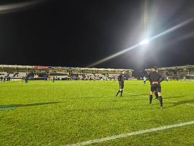 The image displays a football pitch at night, located within Victoria Park (Suit Direct Stadium) in Hartlepool, United Kingdom. The scene is illuminated by artificial floodlights, with a prominent pylon visible on the right emitting a strong light beam. The foreground features a well-maintained green grass pitch with white line markings.
Two individuals, possibly players or officials, stand in the mid-foreground with their backs mostly to the viewer, wearing dark athletic attire. One individual on the right has hands clasped behind their back. Further back on the pitch, several other individuals in dark and light-colored sportswear are sparsely distributed, appearing to be standing or walking.
The background consists of stadium stands with multiple tiers of blue and white seating. A limited number of spectators are visible in these stands. Advertising hoardings run along the front of the stands, displaying text such as "GB.co.uk," "CYMRU KNOWLE & STAND," and "BRUNEL." A white goalpost structure is partially visible in the far right background. The overall environment suggests a sporting event or training session under artificial lighting.