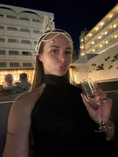 The media file is a photograph taken at night in Es Camp De Mar, Spain. The image features a young woman as the primary subject, who is looking towards the camera with a playful expression, sticking her tongue out slightly. She is wearing a black top and an ornate pearl headband. In her right hand, she holds a small shot glass filled with a light-colored liquid, possibly alcohol. Behind her, a white building with multiple floors and windows is visible, illuminated by warm lights. The sky is dark, indicating it's nighttime. There are other people in the background, including a man wearing a patterned shirt, partially visible. The overall mood appears to be festive and casual, suggesting a social gathering or celebration.