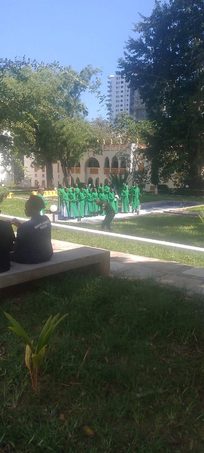 The image displays an outdoor scene in Tudor ward, Kenya, under a clear blue sky. In the lower-left foreground, two individuals are seated on a concrete bench; one wears a dark t-shirt with the white text "HABARI KIUMBANI PEPETA" on the back. A paved pathway separates this area from a manicured green lawn.
In the midground, a group of approximately 25-30 individuals, primarily young adults, stands in organized rows on the grass. They are uniformly dressed in long, flowing green robes over white undergarments. One individual in dark attire, possibly with yellow accents, actively gestures or moves in front of this group, leaning forward with arms outstretched. To the right of the group, a blue-tiled water feature is visible.
In the background, a multi-story cream-colored building with distinctive arched windows and decorative golden-brown architectural details is partially visible through green trees. Above and behind this structure, a tall, modern, light-colored high-rise building is visible against the clear sky. The overall environment features lush green grass and numerous trees.