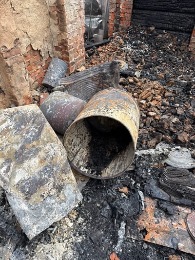 The image depicts the aftermath of a destructive event, consistent with a fire, located in the city of Nedryhailiv, Ukraine. The scene is a cluttered ground-level view of structural wreckage and debris. In the foreground, multiple metal containers, identified as barrels, lie amidst charred material. One barrel, oriented horizontally in the lower-right, is open-ended, displaying internal black residue and external rust and soot. Adjacent to it, a large, fractured slab of scorched material, possibly concrete or stone, rests on the ground. A severely warped and blackened piece of corrugated metal or a similar structural element is also visible among the debris. The ground is covered in a dense layer of black ash, burnt wood fragments, reddish-brown brick rubble, and various unidentifiable charred remnants. In the background to the left, a damaged brick wall with peeling plaster exposes red brickwork. To the right, a section of a structure appears entirely carbonized and black. A distorted window opening, partially covered by translucent sheeting, is visible in the upper-left background. No individuals are present in the frame.