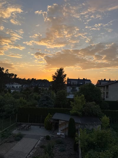 This is an outdoor, full-frame, vertically oriented, high-angle shot of a residential area during sunset in Marki, Poland. The sky is filled with a mix of light blue and orange hues from the setting sun, with scattered clouds tinged with yellow and orange. Below the sky, a line of trees forms a dark silhouette against the bright horizon.
In the mid-ground, several multi-story residential buildings with dark roofs are visible, interspersed with dense green foliage. The buildings appear to be apartment blocks or townhouses.
The foreground shows a backyard area with a paved patio, a picnic table with benches, a small brick shed, and a neatly trimmed hedge. A chain-link fence runs along the left side of the yard, bordering a garden area with some plants. The overall impression is of a quiet suburban or semi-rural setting at dusk.