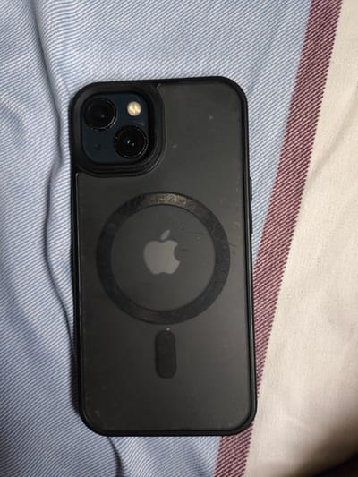 The image shows an iPhone 13 Pro in a clear protective case, lying on a striped fabric surface. The phone is positioned vertically, with the camera array visible at the top. The case has a circular magnetic charging area on the back, with the Apple logo prominently displayed within it. The fabric beneath the phone has alternating blue and white stripes on the left, transitioning to stripes of maroon, white, and blue on the right. The lighting suggests it is daytime, and the setting appears to be indoors, possibly a bed or sofa, given the texture and pattern of the fabric. There is no visible text in the image. The scene is static, with the phone simply resting on the surface, and no activity is taking place. The overall impression is that of a product photograph or a casual shot of a personal item.