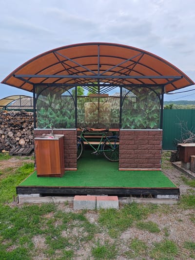 An outdoor gazebo with a curved, orange translucent roof supported by a black metal frame stands on a raised wooden platform covered in artificial grass. The gazebo has decorative green leafy fabric panels on the back and sides, and the lower portion of the back wall is constructed from brown brick-like siding. Inside, a green bicycle leans against wooden benches. To the left of the gazebo, a wooden cabinet with a stainless steel sink and faucet is situated on the artificial grass. To the left of the gazebo, a large pile of cut firewood is stacked on wooden pallets. The ground surrounding the platform is covered in dirt and sparse grass. In the background, a green metal fence separates the property from a forested hillside. The sky above is overcast. The scene is located in the village of Peschanakoye, Russia.