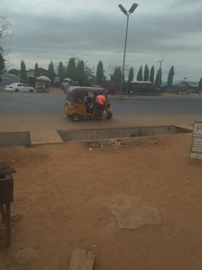 The image shows a yellow three-wheeled vehicle parked on the side of a paved road in Junction, Nigeria. The vehicle has three people inside. The vehicle is parked in front of a concrete curb with a gap that appears to be a drainage ditch. The curb is on the edge of an unpaved area of dirt and gravel. There is a street lamp on the other side of the road. The road is empty, and the sky is overcast. There is a sign for a nearby parking lot.  The scene is likely in an urban area.