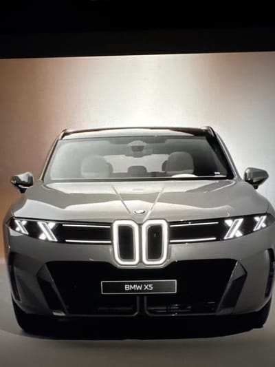 The image shows a frontal view of a grey BMW X5. The car has a distinctive grille with illuminated vertical elements, and sharp, angular headlights. A black license plate with "BMW X5" written in white is visible below the grille. The car is positioned against a gradient background that transitions from a darker hue at the top to a lighter, almost white color at the bottom. The lighting suggests a studio setting or a controlled environment, with the car being the sole focus. There are no people or any urban environmental elements visible in the image, therefore it is impossible to determine its location in Sant'Angelo di Piove di Sacco, Italy.