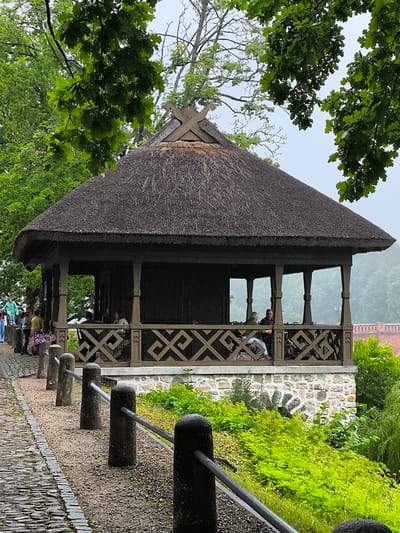 The image depicts a charming thatched-roof gazebo situated in the historic town of Kuldīga, Latvia. The gazebo features intricate wooden lattice railings and is supported by sturdy wooden posts. Inside, a couple is seated at a table, engaged in conversation, while other visitors are visible walking along a cobblestone path to the left of the structure. The gazebo is perched on a stone foundation, surrounded by lush green foliage. The overall atmosphere is tranquil and inviting, suggesting a peaceful day in this picturesque European town.