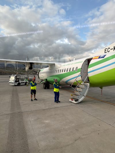 The image captures an active airport tarmac scene in Granadilla de Abona, Spain, during daytime under a partly cloudy sky. The central subject is a turboprop aircraft, possibly a regional airliner, with a distinctive livery featuring green, white, and light blue stripes.
The aircraft's main cabin door is open, revealing integrated airstairs extending to the ground. Red and white striped cones, bearing the text "REMOVE BEFORE FLIGHT," are placed around the bottom of the stairs. Visible text on the fuselage includes "EC-M" near the tail, indicating its registration, and "LINEAS AEREAS DE CANARIAS" along the side. A flag resembling the Spanish national flag with a coat of arms is also seen on the tail section. Inside the open door, the word "Canair" is visible.
Two ground crew members are prominent in the foreground. Both wear high-visibility yellow vests over dark blue attire, with one in shorts and the other in long pants. The crew member closer to the front of the plane stands near a white pushback tug, which has "Better" written on its side. This individual holds what appears to be a marshalling wand or headset. The second crew member stands further back, near the aircraft's engine and propeller, also holding a similar item. A third individual in an orange vest is partially visible behind the second crew member.
The ground is paved concrete or asphalt, showing various markings and textures. In the background, beyond the aircraft's wing, distant mountains are visible under the cloudy sky, possibly reflected in the wing's surface or seen through a window. The scene suggests activities related to aircraft ground operations, such as preparing for departure or servicing upon arrival.