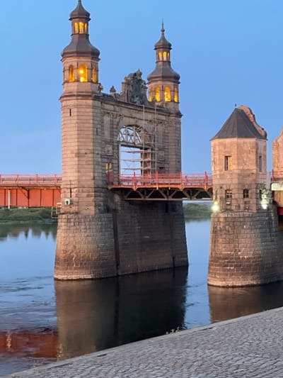 This image shows the Sovetsk Bridge, also known as the Tilsit Bridge, at dusk. The bridge spans a body of water, likely the Neman River, separating the city of Sovetsk, Russia, from the Lithuanian border. The bridge features prominent stone towers with illuminated windows and spires, one of which is undergoing renovation with scaffolding visible. A bright orange bridge section is visible behind the stone structure. The sky is a clear blue, indicative of twilight. The water reflects the bridge and the sky. A cobblestone walkway lines the foreground. There are no people visible in the image.