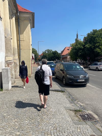 Two people are walking on a cobblestone sidewalk in Stupava, Slovakia. The person in the foreground is wearing a white t-shirt and black shorts, with a backpack. The second person, further down the sidewalk and slightly ahead, is wearing a dark hooded jacket and shorts, and is carrying a red plastic bag.
The scene appears to be a street in a town, with buildings lining one side of the road and a street with parked cars on the other. A dark gray Volkswagen is parked on the street, with a Slovakian license plate reading "MA 369ED". The sky is clear and blue, suggesting a sunny day, likely in the late morning or early afternoon due to the sharp shadows cast on the ground. Trees with lush green foliage are visible, as are buildings with red tile roofs and light yellow walls. A church steeple is visible in the background. The overall atmosphere is one of everyday activity on a pleasant day.