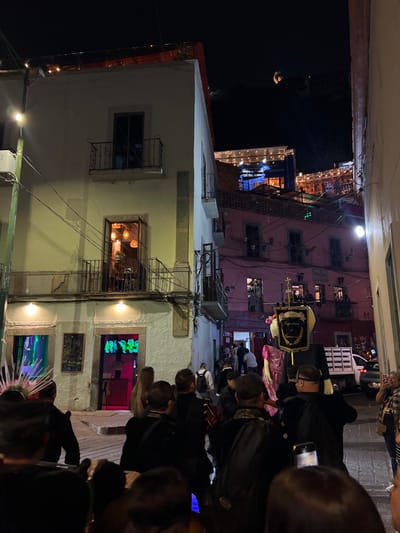 The image captures a nighttime street scene within a narrow callejón in Guanajuato City, Mexico. In the mid-ground, a crowd of approximately twenty individuals, largely dressed in dark clothing, occupies the street. Prominently featured among them are two large mojiganga figures: a male figure dressed in a black suit with a large, elaborate headpiece featuring a black banner with gold trim, an insignia, and a cross, and a partially visible female figure in a pink dress to its left.

To the left of the frame, a multi-story white building features two iron-railed balconies on its upper floors; the lower balcony's door is open, revealing interior lighting from multiple hanging fixtures. Below, at street level, is a commercial establishment with green and pink neon signage spelling "NIRAS" and a small chalkboard sign. A street lamp is mounted on this building. To the right, a multi-story pink building with several balconies extends into the background. Overhead utility wires are visible across the upper portion of the scene. In the far background, on a dark hillside, several illuminated structures with string lights are visible, with the faint outline of the Pípila monument discernible against the night sky. A white pickup truck is partially visible on the far right in the background.