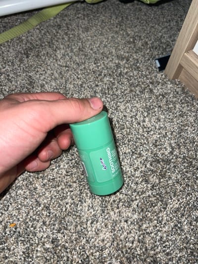A hand holds a green deodorant stick with the brand name "Glow" and text reading "SOOTHING TEA TREE CLAY" on the label. The stick is on a textured carpet.