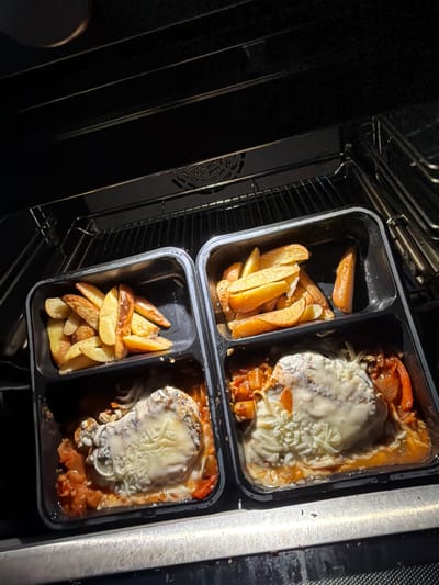 The image shows two black plastic trays placed in an oven. Each tray is divided into two compartments. One compartment in each tray contains potato wedges, while the other compartment contains a food item topped with melted cheese and a tomato-based sauce. The oven interior is visible, showing the rack and a dark interior space. The location is the city of Ede, Netherlands is not discernible from the given image.