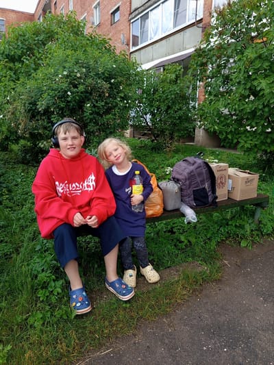 Two children, a boy and a girl, are sitting on a green wooden bench outdoors. The boy, on the left, is wearing a red hoodie, dark pants, and blue crocs. He has headphones on. The girl, on the right, is wearing a dark blue dress and beige crocs. She is holding a bottle of water. Behind them, on the bench, are two cardboard boxes, a grey sleeping bag, a purple backpack, and an orange plastic bag. The setting appears to be in front of a brick building with bushes and trees. The image is likely taken in Gdov, Russia, given the presence of products with Russian labels on the boxes. The ground is covered with grass and asphalt.