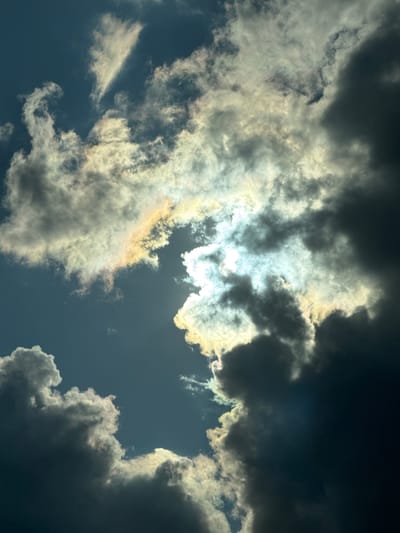 The image presents a dramatic and dynamic sky dominated by an intricate interplay of clouds and sunlight. Dense, dark clouds occupy significant portions of the frame, suggesting a heavily overcast or potentially stormy atmosphere. However, the scene is powerfully illuminated by the sun, which is not directly visible but casts an intense, brilliant glow from behind the central cloud mass. This light filters through the clouds, creating bright, almost ethereal highlights in shades of white, yellow, and subtle iridescent colors along their edges.
Lighter, more translucent cloud formations are also present, some appearing wispy and diffuse, further scattering the sunlight. Patches of the deep blue sky are visible through gaps in the cloud cover, offering a stark contrast to the shadowy, darker cloud bodies. The overall impression is one of natural grandeur and atmospheric power, with the sun's light piercing through heavy clouds. No human figures, man-made objects, or discernible text are visible. The weather appears cloudy with strong sunlight breaking through, likely during daytime in Verkhniy Mamon, Russia.