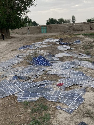 This image captures a rural outdoor scene in Qila Saifullah, Pakistan, under an overcast sky. The foreground and midground are dominated by numerous broken and fragmented solar panels, scattered across dry, dusty ground. These panels, characterized by their blue color and white grid patterns, appear to be extensively damaged, varying in size and shape. Some pieces are partially covered in dust or dirt.

On the left side of the frame, a large, mature tree with dense green foliage stands prominently. Two young individuals, likely children, are visible near the base of this tree, looking towards the right side of the frame. In the mid-ground, a person is standing near a low, light-brown mud-brick wall that extends horizontally. This wall encloses part of the area, and features a distinctive light-colored, possibly green, double door set within it. Another person is partially visible near the right edge of the background, closer to other mud-brick structures.

The background is composed of traditional mud-brick buildings and walls, suggesting a village or rural settlement. The ground is largely unpaved and dusty, with sparse patches of green vegetation or weeds growing amongst the debris. The diffuse lighting indicates an overcast day, possibly morning or late afternoon. The scene conveys a sense of neglect or the aftermath of an event, with the broken solar panels lying strewn about. No discernible text is visible in the image.