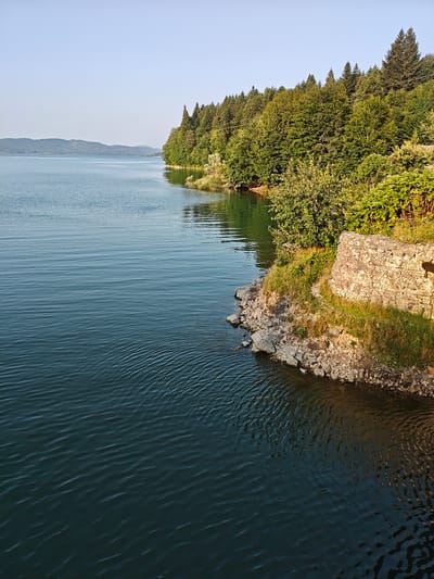 The image depicts a tranquil lake scene with lush green trees lining the shore. The water is a deep blue, with gentle ripples creating a mesmerizing pattern. In the foreground, a rocky embankment is visible, interspersed with patches of grass and a weathered stone structure that appears to be part of a wall or fortification. The trees on the bank are a mix of deciduous and coniferous species, their leaves rustling in the soft breeze. In the distance, rolling hills fade into a hazy horizon under a clear blue sky. The overall impression is one of serene natural beauty, possibly a reservoir or a large natural lake. The location is identified as Kharistvala, Georgia, suggesting a picturesque natural setting within the country. There are no people or animals visible in the scene, emphasizing the solitude and peacefulness of the environment. The lighting suggests it is either early morning or late afternoon, with the sun casting long shadows and illuminating the landscape with a warm, golden hue.