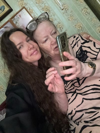 The image shows two women, likely mother and daughter, taking a selfie in a room. The woman on the left has long, dark, wavy hair and is wearing a black top. The woman on the right, who appears to be older, is wearing glasses and a tan and black striped top. She is holding a smartphone and taking the photo. The background appears to be a wall with a floral wallpaper. The room seems to be a living room or bedroom, with framed pictures on the wall. The overall impression is an intimate and casual moment captured between two family members. There is no specific information in the image to suggest a location in Turinsk, Russia.