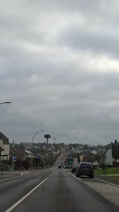 The scene depicts an upward-sloping street in Frisange, Luxembourg, under a uniformly overcast sky. The asphalt road surface is dark and visibly wet. A solid white line delineates the left edge of the road, while a broken white line marks the center for opposing traffic. On the immediate left, a portion of a light-colored building with a dark roof is visible, alongside a public transport shelter with a green waste bin. A dark street lamp arm extends into the frame from the upper left.
On the right side of the foreground, a dark-colored SUV, consistent with a BMW X5, is parked facing uphill on a cobbled shoulder adjacent to a grassy embankment. Further up the road, a green traffic light is visible on the right side. Multiple vehicles are present on the roadway, with several cars observed driving uphill in the distance.
The mid-ground and background feature a residential area with numerous houses characterized by light-colored facades and dark, pitched roofs, built into the rising terrain. Deciduous trees and shrubs, largely bare, are interspersed among the structures. Dominating the distant hilltop skyline is a prominent, multi-level concrete tower, likely a water tower, with a dark, enclosed upper section that displays what appears to be scaffolding or structural modifications. A yellow construction crane is positioned to the right of this tower. A slender, light-colored church spire is faintly visible on the distant left horizon, rising above the surrounding buildings. No individuals are discernible in the image.